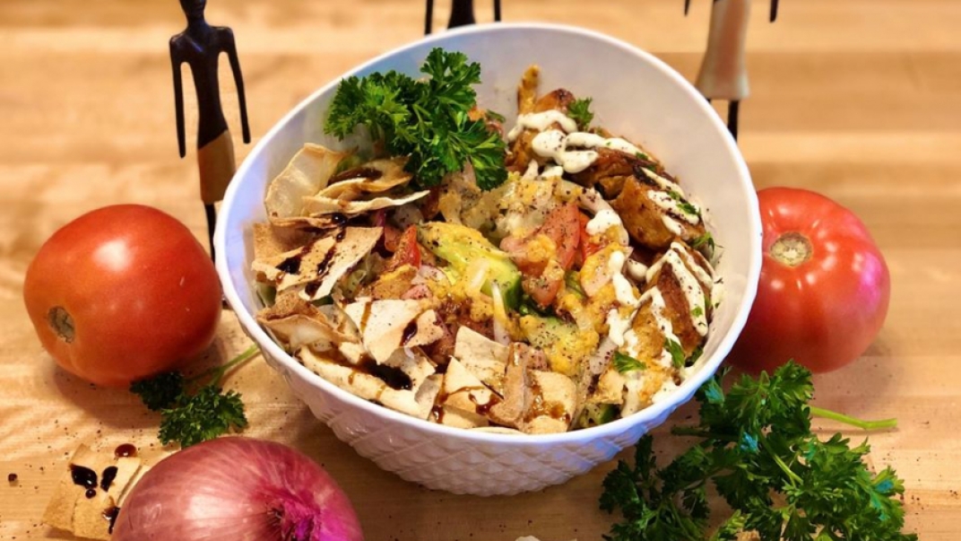 A bowl of food with pita chips crumbled into the bowl, cilantro, and meat from a kebab drizzled with sauce. There are whole tomatoes and an onion around the bowl.