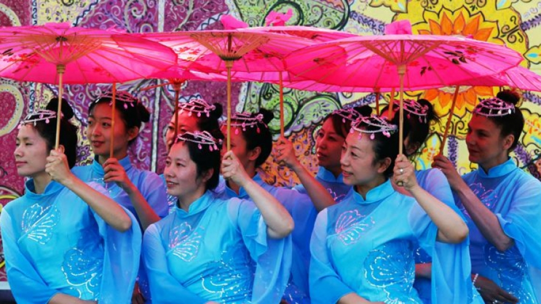 Several Asian women wearing light blue dresses holding up red umbrellas