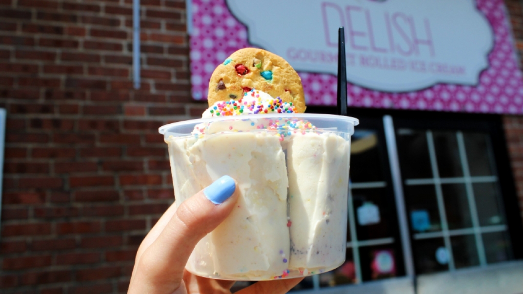 A hand holding a cup of vanilla ice-cream with a cookie on top in front of the Delish store sign. 