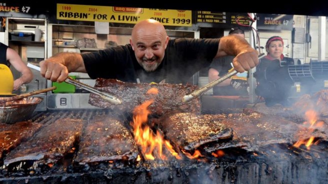 Man standing over a grill with a lot of ribs on it, holding a large rib