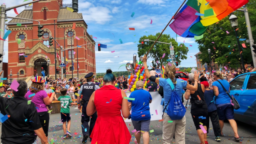 Many people walking down the street of Fredericton with confetti in the air and pride flags