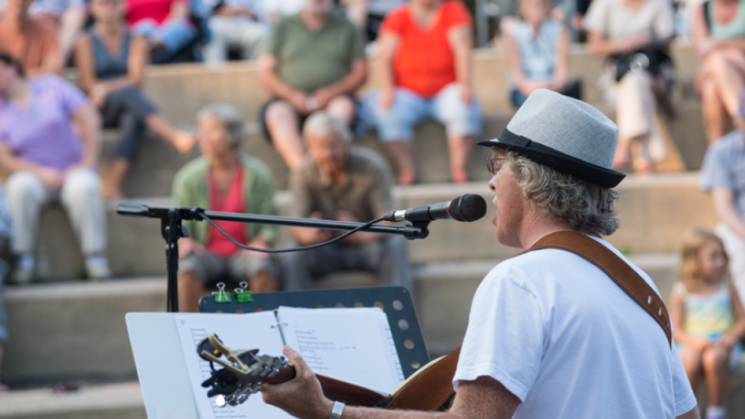 shot of a man playing a guitar and singing with a music stand and sheets in front of him with people sitting on outdoor steps in the background