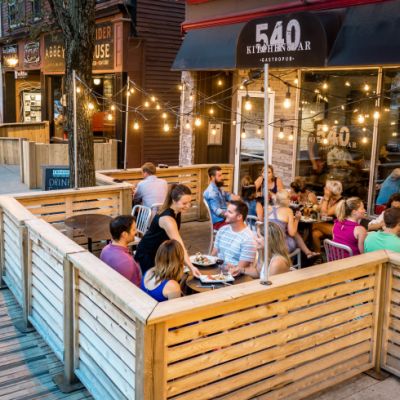 A wood patio built on a side walk. The has 4 full tables and the black awning above it reals 540 Kitchen & Bar