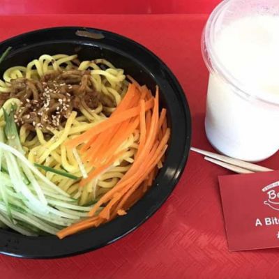 A red cafeteria tray with a black take out container of noodles and julienned cucumbers and carrots on it. Also on the tray is a to go cup holding a white liquid, chopsticks and a business card.