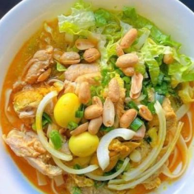 A white bowl with a yellow broth dish, featuring leafy greens, nuts, and onions.