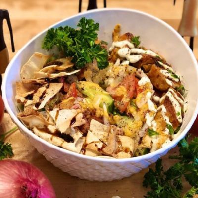 A bowl of food with pita chips crumbled into the bowl, cilantro, and meat from a kebab drizzled with sauce. There are whole tomatoes and an onion around the bowl. 