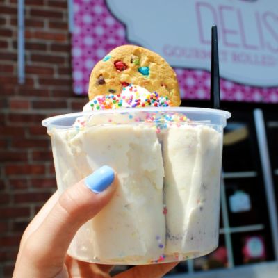 A hand holding a cup of vanilla ice-cream with a cookie on top in front of the Delish store sign. 