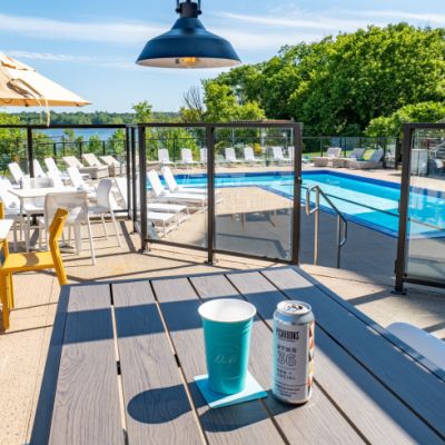 A sunny pool side. The pool is surrounded by white lounge chairs. Up a level is the dining area, with a table with a drink can on it.