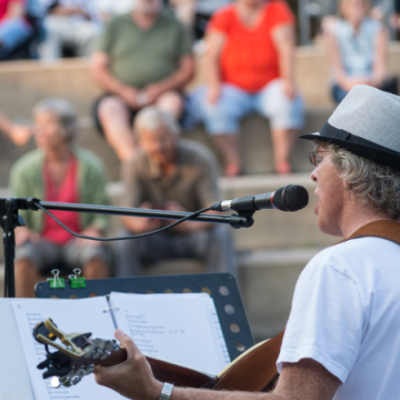 shot of a man playing a guitar and singing with a music stand and sheets in front of him with people sitting on outdoor steps in the background