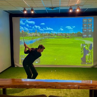 Man swinging golf club at golf ball. There is an electronic golfing projection on a screen in the background