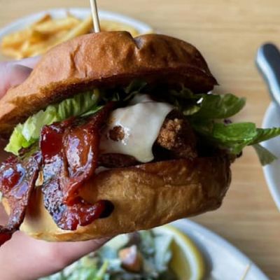 Someone holding a bacon cheeseburger close to a camera with burgers and fries on the table in the background