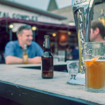 Bartop with beer bottle and pint on them. There are people conversing in the background.
