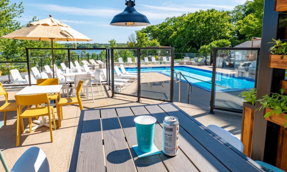 A sunny pool side. The pool is surrounded by white lounge chairs. Up a level is the dining area, with a table with a drink can on it.
