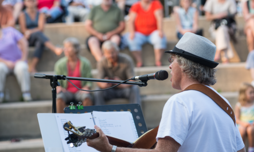 shot of a man playing a guitar and singing with a music stand and sheets in front of him with people sitting on outdoor steps in the background