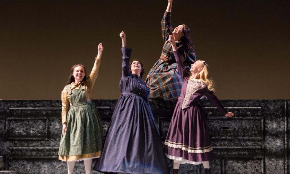 four women in period clothing raise their fist on a theatre stage